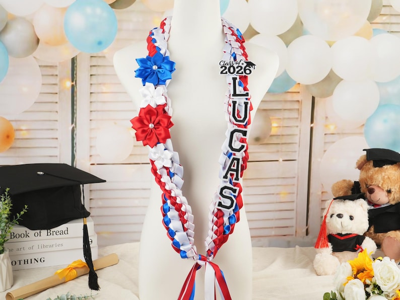 May include: A graduation lei in red, white, and blue ribbon, adorned with blue and red flower accents. The lei features the name "LUCAS" and "Class of 2026" on a black background. A festive graduation decoration.