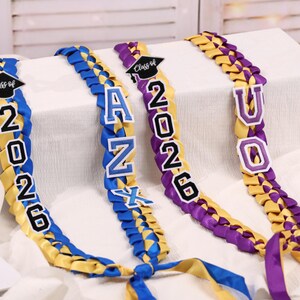 May include: Braided graduation leis in yellow, blue, and purple. Each lei features the year "2026" and a graduation cap, with additional letters spelling out a name. The leis are displayed on a white surface.