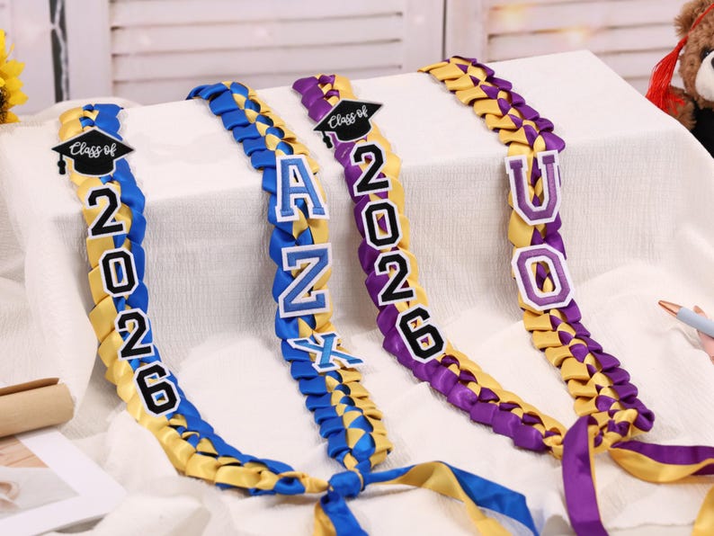 May include: Braided graduation leis in yellow, blue, and purple. Each lei features the year "2026" and a graduation cap, with additional letters spelling out a name. The leis are displayed on a white surface.