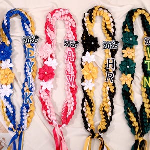 May include: Four graduation leis in blue, pink, black, and green, each with a different name spelled out in block letters. Each lei has a "Class of 2026" tag and is decorated with fabric flowers and ribbons.