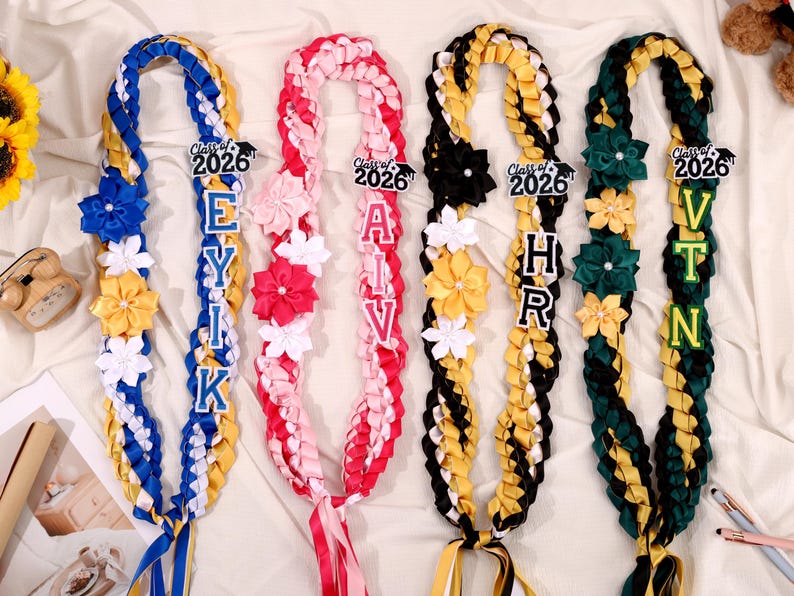 May include: Four graduation leis in blue, pink, black, and green, each with a different name spelled out in block letters. Each lei has a "Class of 2026" tag and is decorated with fabric flowers and ribbons.
