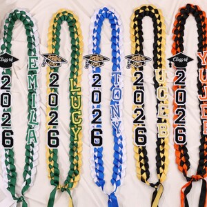 May include: Braided graduation leis in green, white, yellow, blue, black, and orange. Each lei features a graduation cap and the year 2026, along with a name. The leis are a celebratory item for graduates.