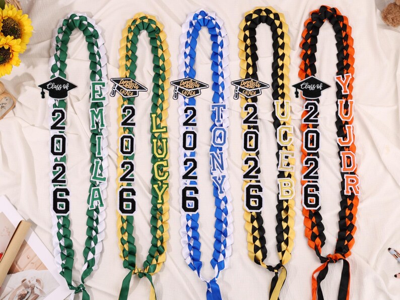 May include: Braided graduation leis in green, white, yellow, blue, black, and orange. Each lei features a graduation cap and the year 2026, along with a name. The leis are a celebratory item for graduates.