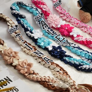May include: Braided graduation leis in various colors, including brown, blue, and pink. Each lei features a name and the year "2026" with decorative flower accents. A teddy bear is visible in the background.