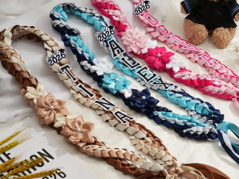 May include: Braided graduation leis in various colors, including brown, blue, and pink. Each lei features a name and the year "2026" with decorative flower accents. A teddy bear is visible in the background.