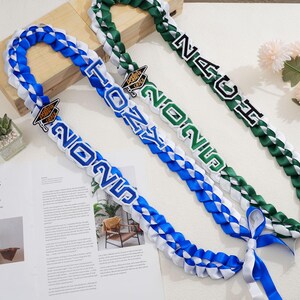 May include: Braided graduation leis in blue, white, and green. Each lei features the year "2025" and a graduation cap charm. One lei reads "TONY", and another reads "NAU". Ribbons are tied at the ends.