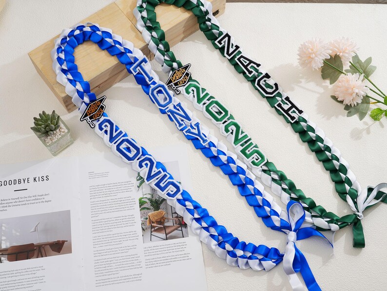 May include: Braided graduation leis in blue, white, and green. Each lei features the year "2025" and a graduation cap charm. One lei reads "TONY", and another reads "NAU". Ribbons are tied at the ends.