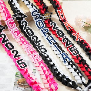 May include: Braided graduation leis in pink, black, white, and red. Each lei features a name and the year "2025" in bold lettering. Some leis include a graduation cap emblem, perfect for celebrating the Class of 2025.