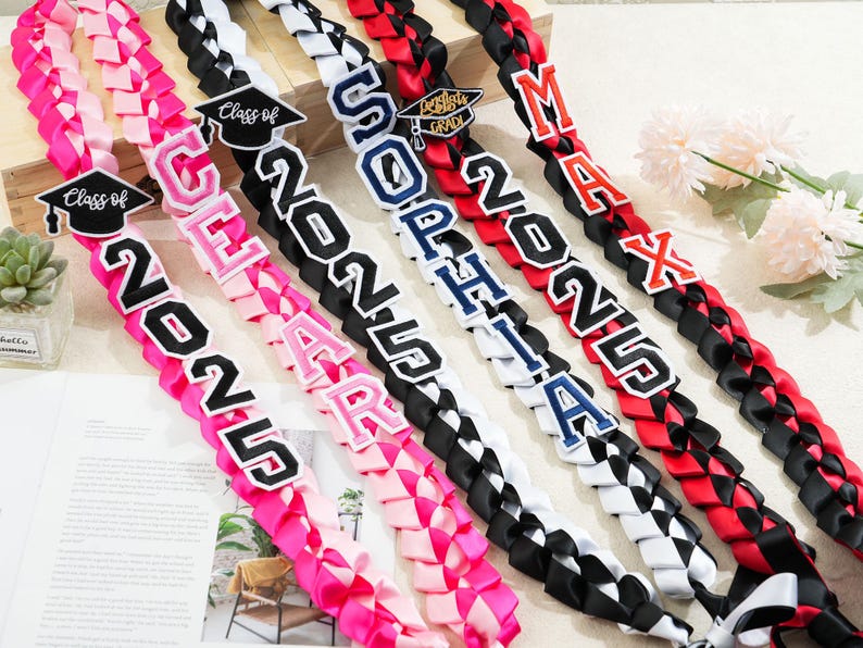 May include: Braided graduation leis in pink, black, white, and red. Each lei features a name and the year "2025" in bold lettering. Some leis include a graduation cap emblem, perfect for celebrating the Class of 2025.