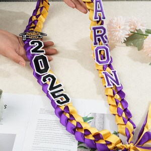 May include: A graduation lei in purple and gold, braided with satin ribbons. The lei features the name "Aaron" in white letters with purple trim, and the year "2025". A small graduation cap charm is also included.