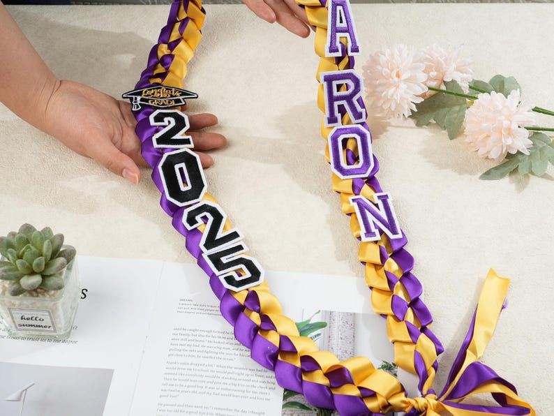 May include: A graduation lei in purple and gold, braided with satin ribbons. The lei features the name "Aaron" in white letters with purple trim, and the year "2025". A small graduation cap charm is also included.