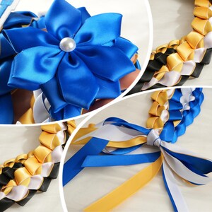 May include: A close-up of a graduation lei featuring a large blue satin flower with a pearl center. The lei is made of woven ribbons in blue, gold, black, and white. The ribbons are braided together to form the lei.
