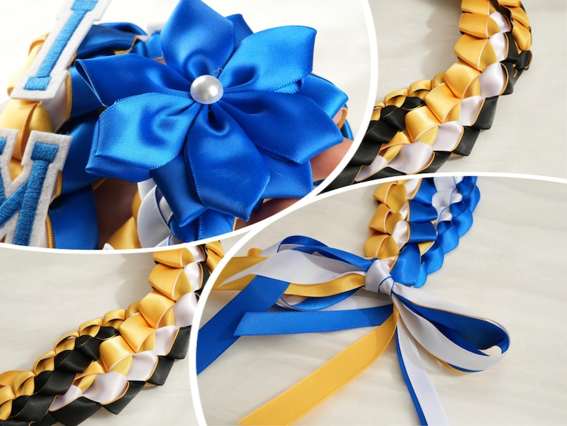 May include: A close-up of a graduation lei featuring a large blue satin flower with a pearl center. The lei is made of woven ribbons in blue, gold, black, and white. The ribbons are braided together to form the lei.