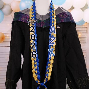 May include: A graduation lei with blue and gold braided ribbons. The lei features the year "2026" and the letters "ANX" in white block letters. A small graduation cap charm is at the top. The lei is displayed on a black graduation gown.