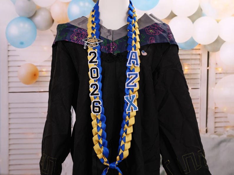 May include: A graduation lei with blue and gold braided ribbons. The lei features the year "2026" and the letters "ANX" in white block letters. A small graduation cap charm is at the top. The lei is displayed on a black graduation gown.