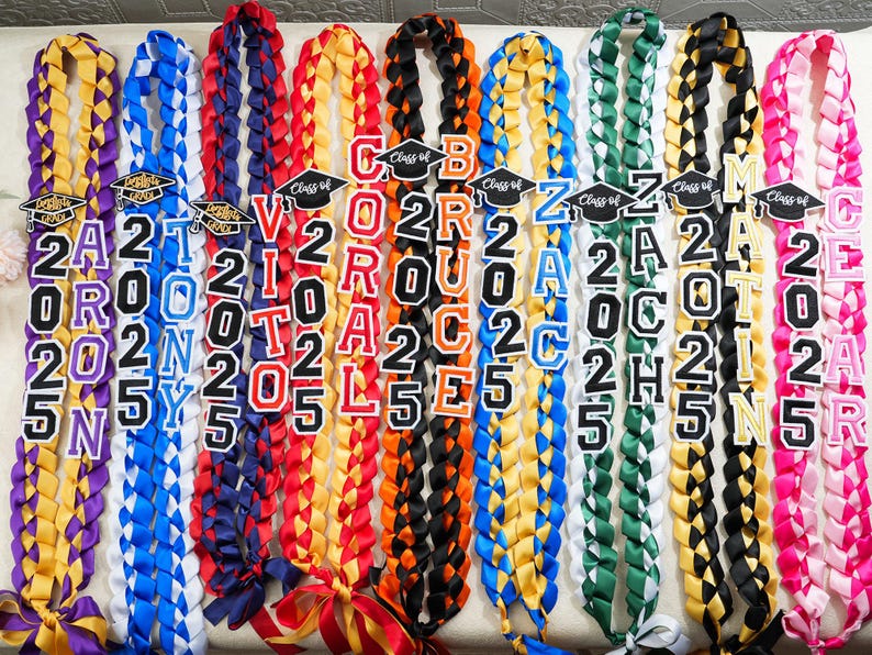 May include: A collection of braided graduation leis in various colors, including yellow, blue, red, and black. Each lei features a name and the year "2025" in bold letters, with a graduation cap emblem. Ribbons are tied at the bottom.