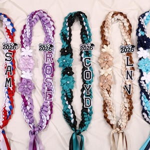 May include: Braided graduation leis in red, white, blue, purple, teal, and brown. Each lei features a name and the text "Class of 2026". Decorated with ribbon flowers and bows, these leis are a celebratory graduation accessory.