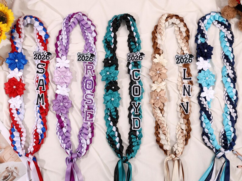 May include: Braided graduation leis in red, white, blue, purple, teal, and brown. Each lei features a name and the text "Class of 2026". Decorated with ribbon flowers and bows, these leis are a celebratory graduation accessory.