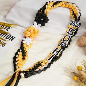 May include: A graduation lei in black, yellow, and white, with the name "SOPHIA" and "Class of 2026" displayed. The lei is decorated with fabric flowers and ribbons. A teddy bear is in the background.