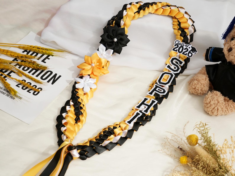 May include: A graduation lei in black, yellow, and white, with the name "SOPHIA" and "Class of 2026" displayed. The lei is decorated with fabric flowers and ribbons. A teddy bear is in the background.