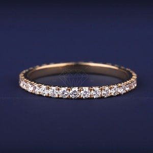 May include: A gold band ring with a continuous row of small, round, clear gemstones. The ring is set against a dark blue background. The ring is a delicate piece of jewelry.