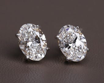 May include: A pair of oval-cut diamond stud earrings. The diamonds are set in a silver metal and sparkle brightly. The earrings are displayed on a brown surface. The image is well-lit, showcasing the clarity and brilliance of the gemstones.