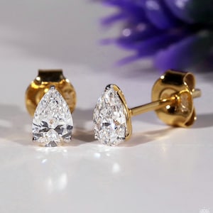 May include: A pair of gold stud earrings featuring pear-shaped diamonds. The diamonds are clear and faceted, reflecting light. The earrings have a classic design with a gold post and backing. The background is a soft white with a blurred purple flower.