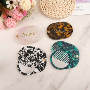 May include: Four decorative compact mirrors with combs. One is pearlescent with the name "Annie", another is tortoiseshell with "Nahum", and the other two have black and white, and green and black patterns. Each has a gold-coloured rivet.