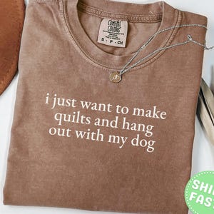Puede incluir: Una camiseta marrón de manga corta con el texto blanco "i just want to make quilts and hang out with my dog" impreso en la parte delantera. La camiseta cuenta con un cuello redondo y una etiqueta de la marca Comfort Colors en el cuello.