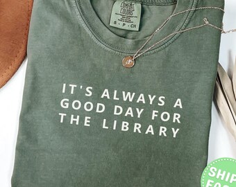 May include: A moss green short-sleeve t-shirt with a crew neckline and the white text "IT'S ALWAYS A GOOD DAY FOR THE LIBRARY" printed on the front. A delicate gold-toned chain necklace with a small circular pendant rests on the shirt collar.