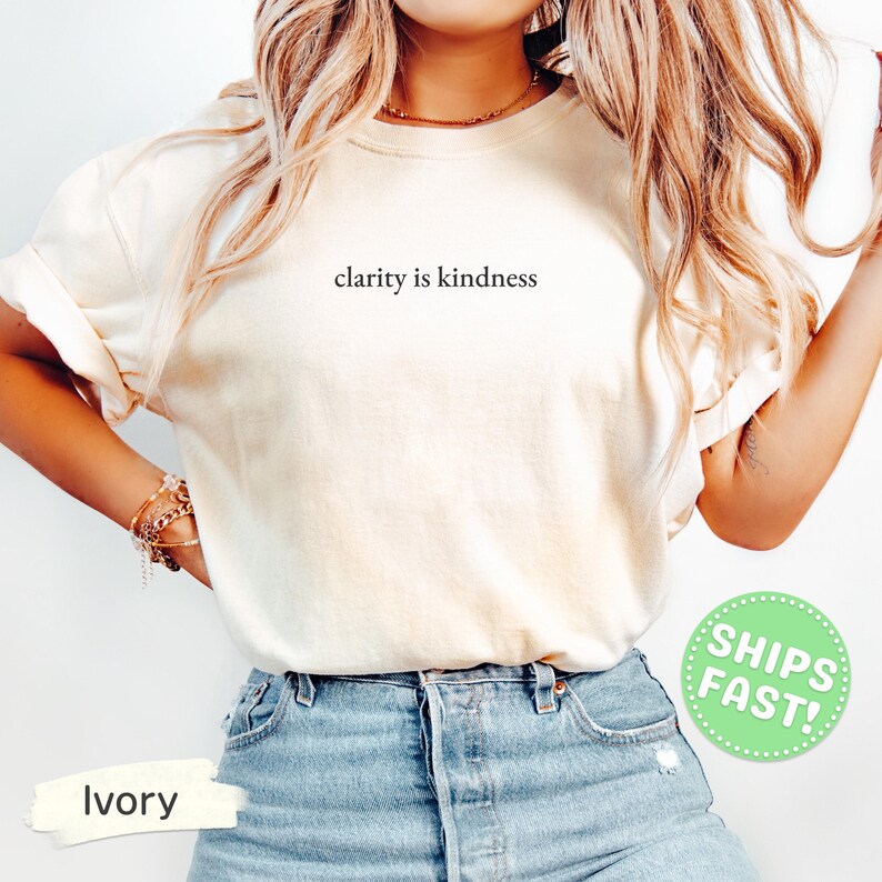 Może przedstawiać: Koszulka z kr&oacute;tkim rękawem w kolorze kości słoniowej z czarnym napisem "clarity is kindness" nadrukowanym na środku klatki piersiowej. T-shirt ma okrągły dekolt i jest zestawiony z jasnoniebieskimi dżinsami, tworząc swobodny wygląd.