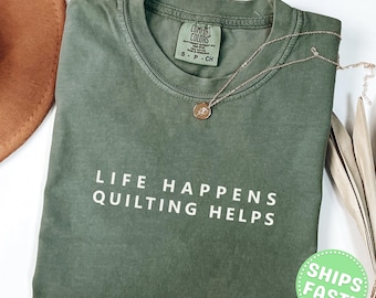 Może przedstawiać: Koszulka z okrągłym dekoltem w kolorze mchu z białym napisem LIFE HAPPENS QUILTING HELPS z przodu. T-shirt posiada metkę marki Comfort Colors przy wewnętrznym dekolcie i jest prezentowany wraz z delikatnym złotym naszyjnikiem na łańcuszku.