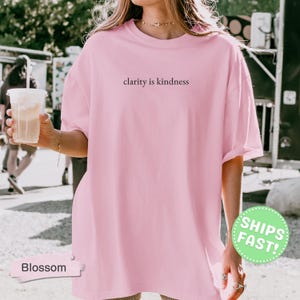Może przedstawiać: Jasnor&oacute;żowy t-shirt z kr&oacute;tkim rękawem z napisem clarity is kindness nadrukowanym prostą czarną czcionką na środku klatki piersiowej. Odzież posiada okrągły dekolt oraz luźny, swobodny kr&oacute;j, co zapewnia codzienny, niezobowiązujący wygląd.