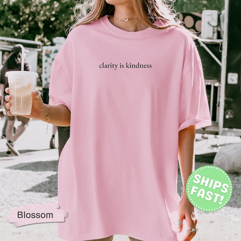 Może przedstawiać: Jasnor&oacute;żowy t-shirt z kr&oacute;tkim rękawem z napisem clarity is kindness nadrukowanym prostą czarną czcionką na środku klatki piersiowej. Odzież posiada okrągły dekolt oraz luźny, swobodny kr&oacute;j, co zapewnia codzienny, niezobowiązujący wygląd.