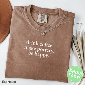 May include: A brown short-sleeved t-shirt with the white printed text "drink coffee. make pottery. be happy." The shirt features a Comfort Colors brand label at the inner neckline and is paired with a delicate silver-toned chain necklace.