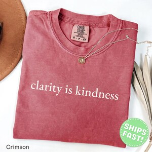 Może przedstawiać: Karmazynowy t-shirt z kr&oacute;tkim rękawem i okrągłym dekoltem, z białym napisem clarity is kindness nadrukowanym na klatce piersiowej. Koszulka posiada metkę marki Comfort Colors przy wewnętrznym kołnierzu i jest zestawiona z cienkim złotym naszyjnikiem.