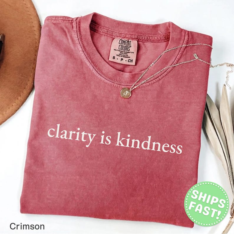 Może przedstawiać: Karmazynowy t-shirt z kr&oacute;tkim rękawem i okrągłym dekoltem, z białym napisem clarity is kindness nadrukowanym na klatce piersiowej. Koszulka posiada metkę marki Comfort Colors przy wewnętrznym kołnierzu i jest zestawiona z cienkim złotym naszyjnikiem.