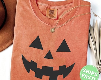Może przedstawiać: T-shirt z okrągłym dekoltem w kolorze wypalonej pomarańczy z czarną grafiką twarzy dyni na przodzie. Koszulka posiada metkę marki Comfort Colors przy wewnętrznym dekolcie. Na tkaninie koszulki spoczywa cienki, złoty łańcuszek.