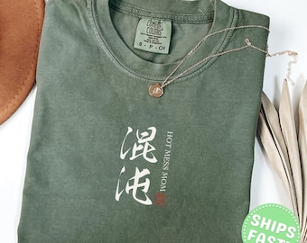 Może przedstawiać: Mchowo-zielony t-shirt z krótkim rękawem z białym napisem "HOT MESS MOM" oraz dużymi białymi japońskimi znakami. Koszulka ma okrągły dekolt z metką marki Comfort Colors wewnątrz i jest zestawiona z delikatnym naszyjnikiem w złotym odcieniu.