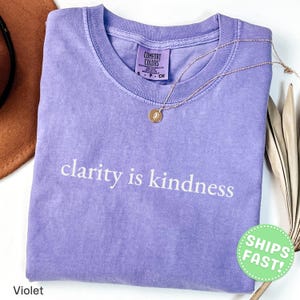 Może przedstawiać: Fioletowy t-shirt z kr&oacute;tkim rękawem, okrągłym dekoltem i spraną fakturą. Napis 'clarity is kindness' jest nadrukowany na piersi w kolorze białym. Złoty naszyjnik z małym okrągłym wisiorkiem spoczywa w okolicach kołnierza koszulki.