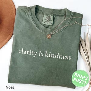 Może przedstawiać: Koszulka z kr&oacute;tkim rękawem w kolorze mchu z białym napisem clarity is kindness nadrukowanym na klatce piersiowej. T-shirt posiada okrągły dekolt oraz metkę marki Comfort Colors wewnątrz kołnierza. Na tkaninie leży cienki złoty łańcuszek.