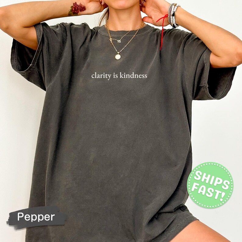 Może przedstawiać: Ciemnoszary t-shirt z kr&oacute;tkim rękawem z nadrukowanym białym napisem clarity is kindness na środku klatki piersiowej. Odzież ma luźny kr&oacute;j z okrągłym dekoltem i wyblakły wygląd w stylu vintage.