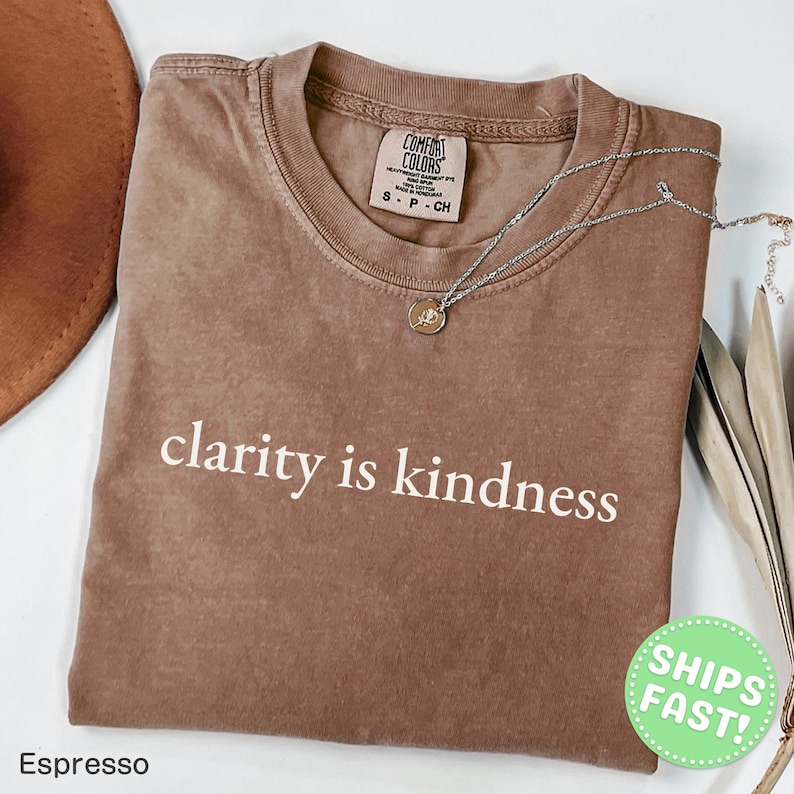 Może przedstawiać: Brązowy t-shirt z kr&oacute;tkim rękawem, okrągłym dekoltem i nadrukowanym białym napisem clarity is kindness na klatce piersiowej. Koszulka posiada metkę marki Comfort Colors przy wewnętrznej stronie kołnierza i jest zestawiona z cienkim srebrnym łańcuszkiem.
