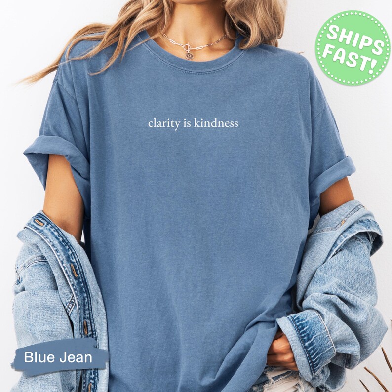 Może przedstawiać: Niebieski t-shirt z kr&oacute;tkim rękawem i okrągłym dekoltem, z białym napisem clarity is kindness na piersi. Koszulka jest noszona z jasnoniebieską kurtką dżinsową zarzuconą na ramiona, widoczny jest r&oacute;wnież srebrny naszyjnik z łańcuszkiem.