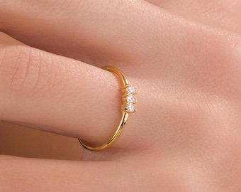 May include: A delicate gold ring featuring three small, clear gemstones. The ring is thin and simple, with the gemstones set in a row. The ring is shown on a person's finger, highlighting its elegant design.