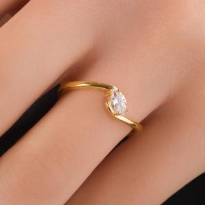 May include: A gold-colored ring with a marquise-cut, clear gemstone. The ring's band curves gracefully, with the gemstone set at an angle. The ring is on a person's finger, with the skin tone visible in the background.