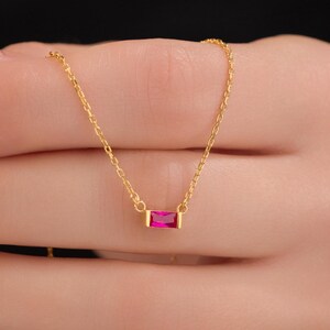 May include: A delicate gold necklace featuring a small, rectangular, bright pink gemstone pendant. The pendant is set in a gold bezel and hangs from a fine gold chain. The necklace is shown against a blurred background.
