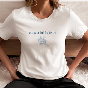 Puede incluir: Camiseta blanca con el texto "cuttest bride to be" y una pequeña ilustración azul de un ramo de flores.