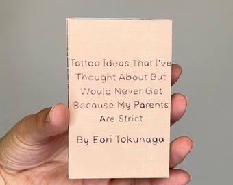 May include: A small, rectangular card with the text "Tattoo Ideas That I've Thought About But Would Never Get Because My Parents Are Strict By Eori Tokunaga" printed in black. The card is held in a hand.