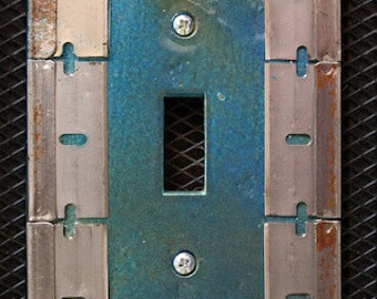 May include: A light switch plate made of metal with a blue and green patina. The plate is surrounded by a frame of metal strips. The plate has two screws and a rectangular opening for the switch.